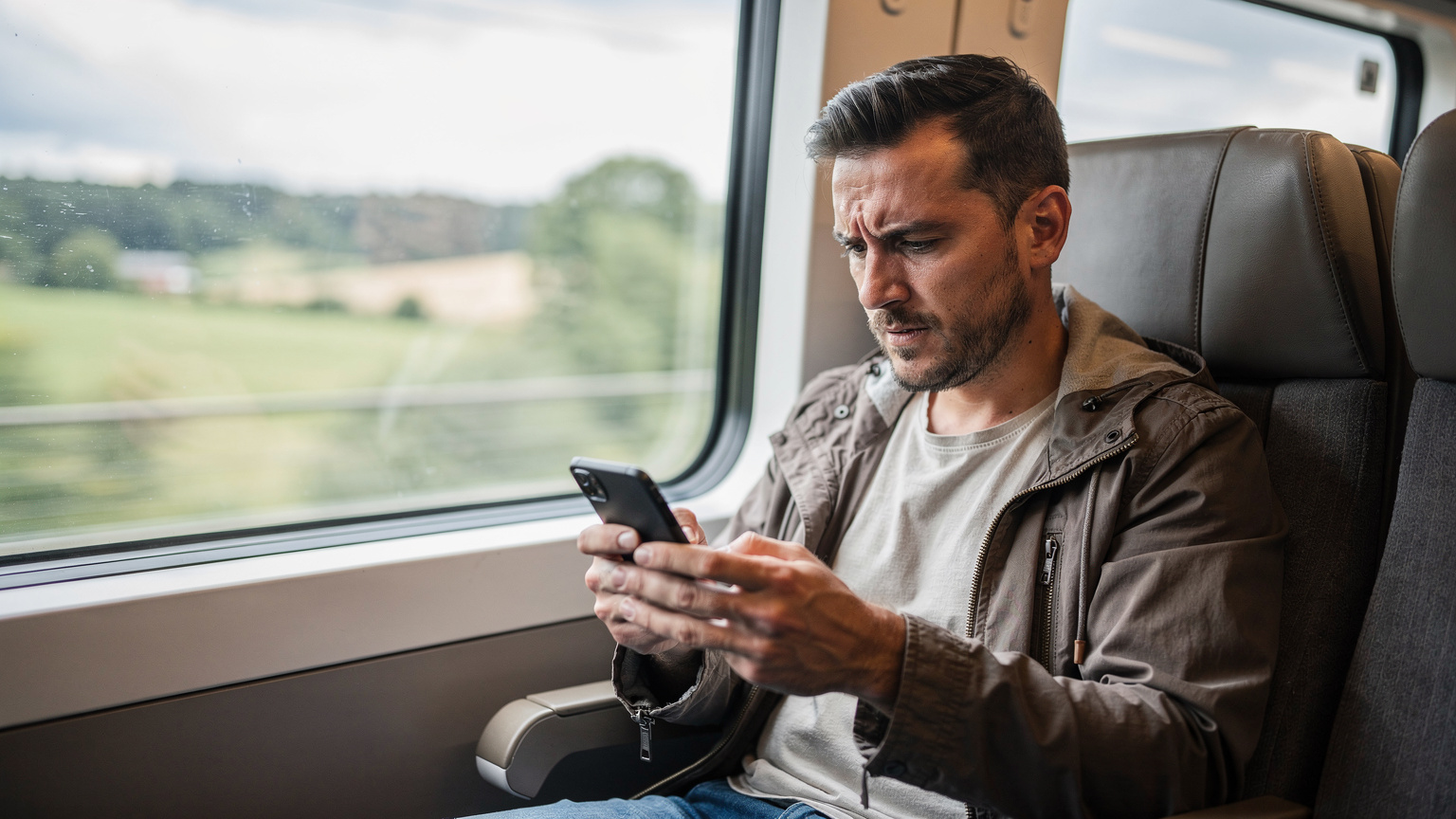 A traveler with a look of mild frustration gazes at their phone screen, which displays a 'No Service' message, while sitting on a high-speed train traveling through the European countryside.