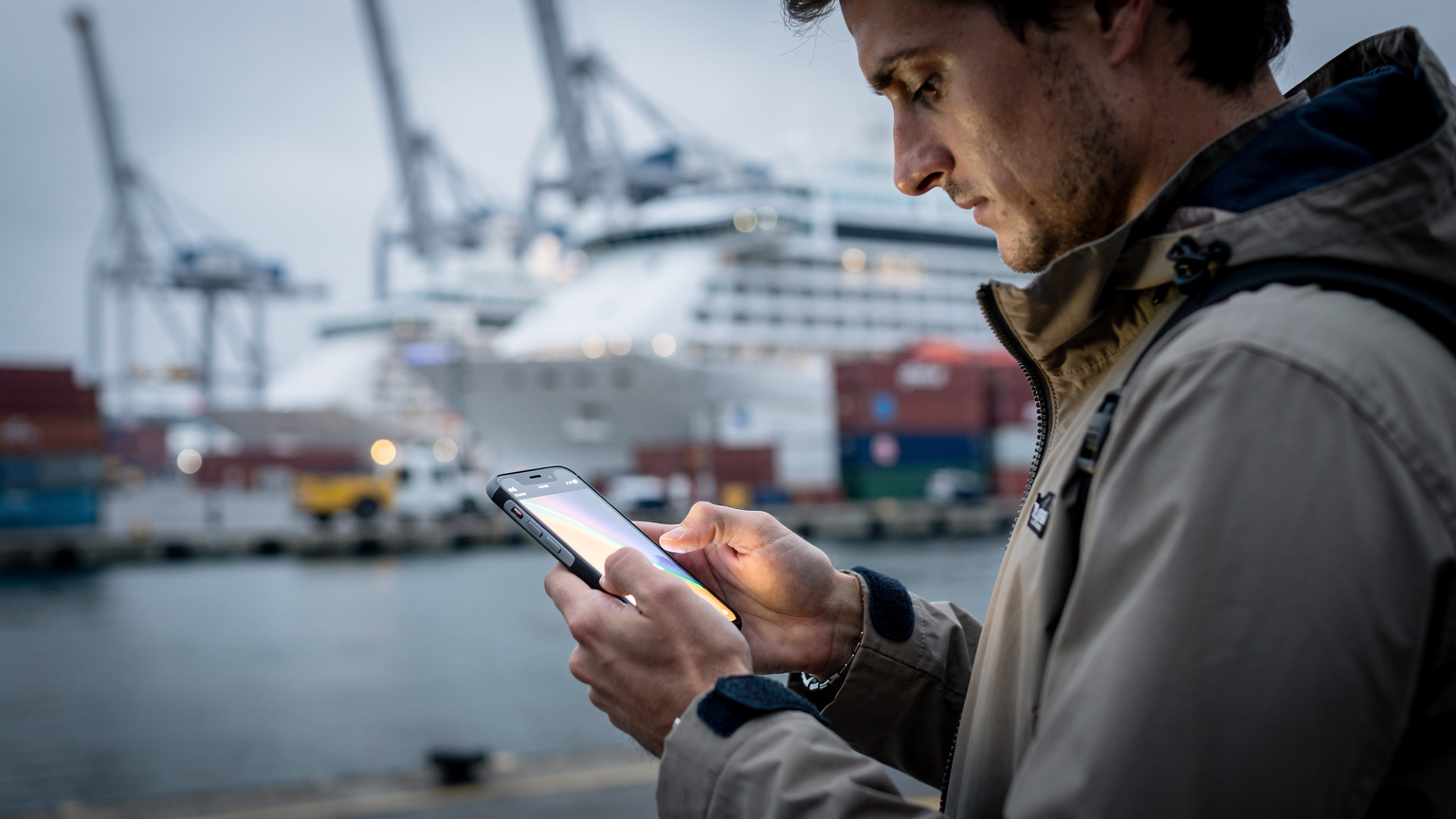 Une personne utilisant son smartphone pour consulter des informations, avec en arrière-plan flou un port de croisière animé avec des bateaux et des grues de chargement, symbolisant le début ou la fin d'une escale.